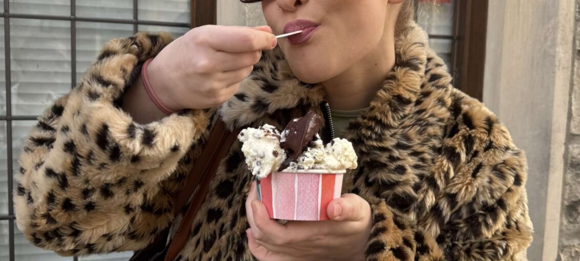 A woman eating a large tub of multi-flavoured ice cream with a slick back bun, wearing a leopard print coat and sunglasses to match, a navy bag is on her shoulder