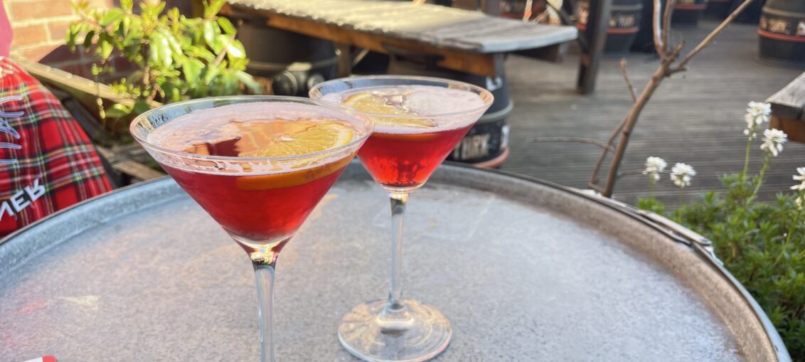 Two Cosmo cocktails in martini glasses with orange slices in them, on table in beer garden.