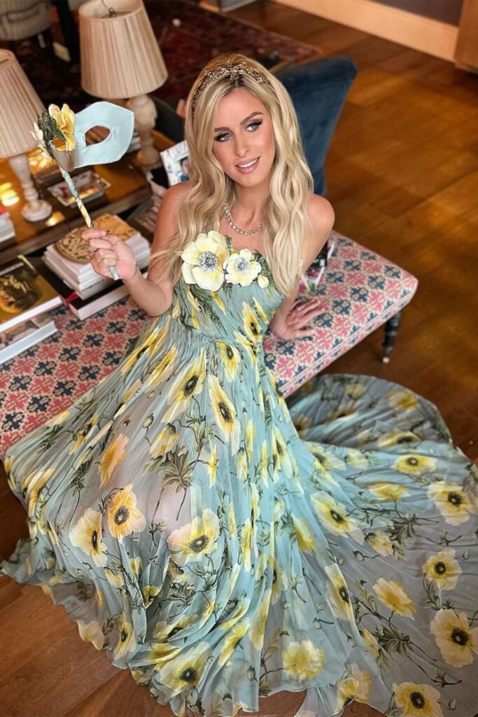 Nicky Hilton wearing a long blue floral dress, one hand resting on a chaise lounge, the other holding up a matching mask. True Italian garden party aesthetics