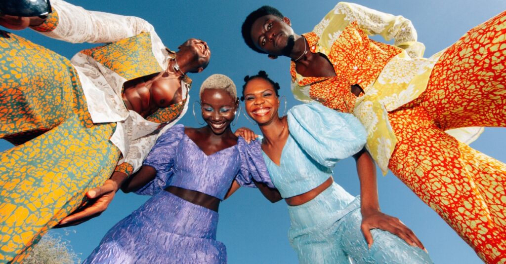 4 people looking down at the camera, each wearing bright clothing, the two women in the middle are laughing, one of the men is stood up and not looking at the camera with a smile on his face and the 2nd man looking down at the camera with a serious look. Dopamine aesthetics to the core