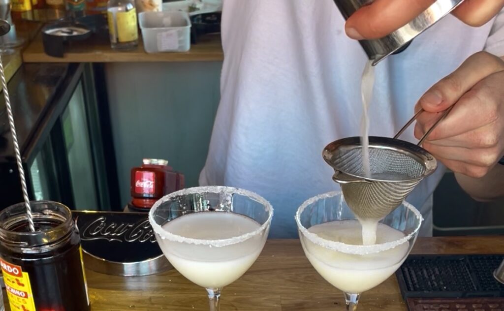 two glasses with sugar rim and lemondrop cocktail inside and a man straining some cocktail from a tin into one of the glasses