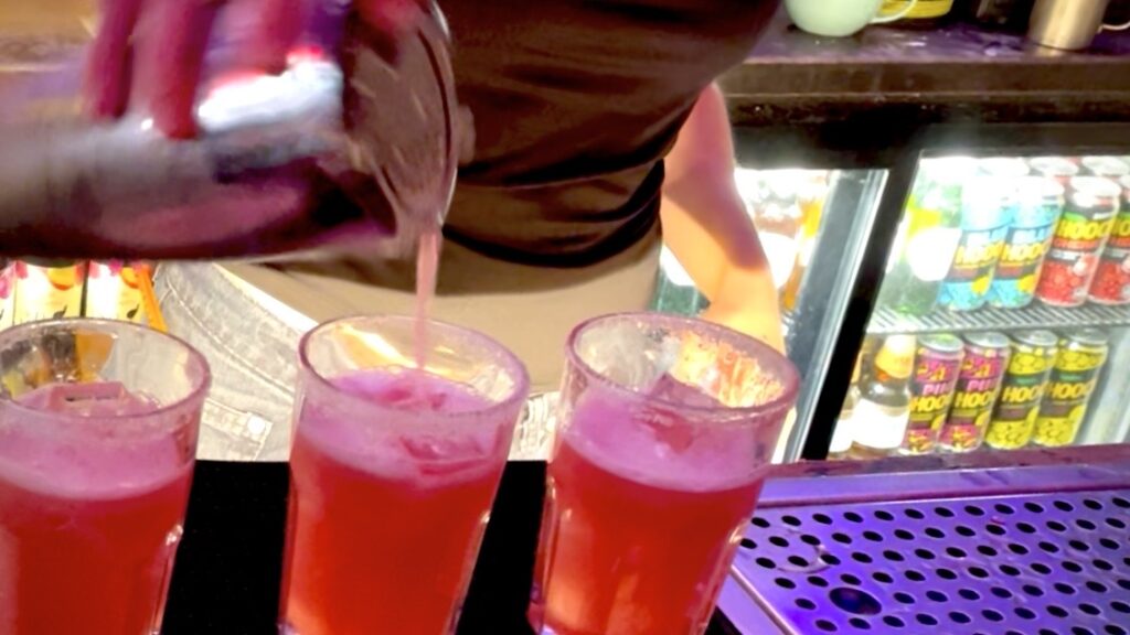 Three glasses with sugar rims, filled with paloma cocktail and bartender pouring some into one of the glasses through strainer.