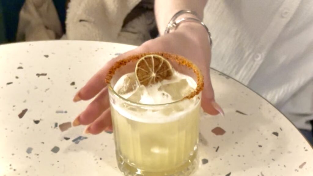 hand reaching for spicy mango margharita with a crystalised orange on top of drink and a chilli rim